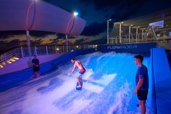 WN, Wonder of the Seas, Brand shoot, man riding boogie board in FlowRider at night, two attendants looking on, fun, action, late sunset,